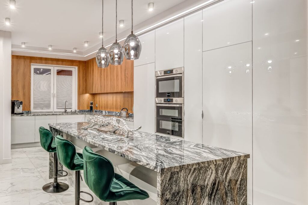 Nowoczesna kuchnia z marmurową wyspą, zielonymi krzesłami i białymi szafkami, oświetlona designerskimi lampami.