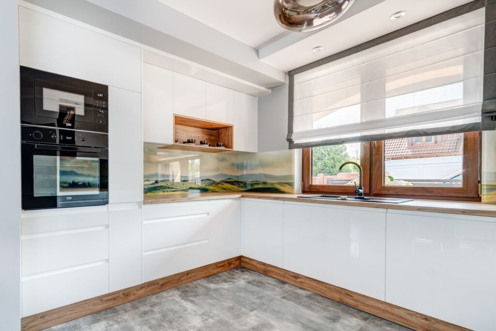 Nowoczesna kuchnia z białymi szafkami, drewnianymi blatami i dużym oknem z widokiem na ogród.