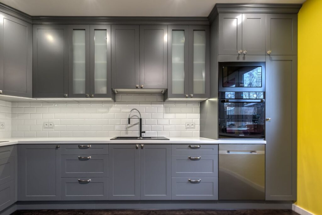Szare szafki kuchenne, białe płytki cegiełki, nowoczesny piekarnik, żółta ściana, ciemna podłoga, zlewozmywak na blacie.