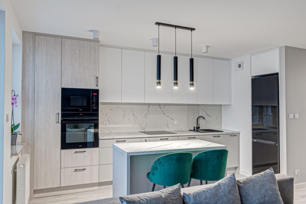 Nowoczesna kuchnia z białymi szafkami, marmurowymi blatami, wyspą i zielonymi krzesłami, oświetlona trzema lampami.