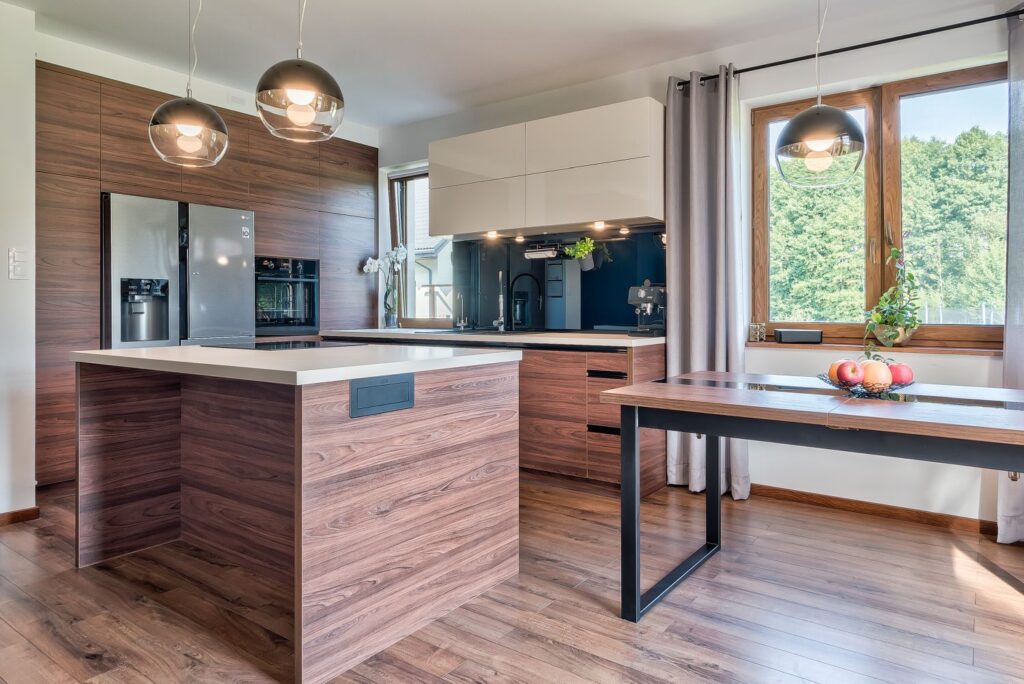 Nowoczesna kuchnia z drewnianymi szafkami, dużymi oknami i wyspą na środku, oświetlona lampami wiszącymi.