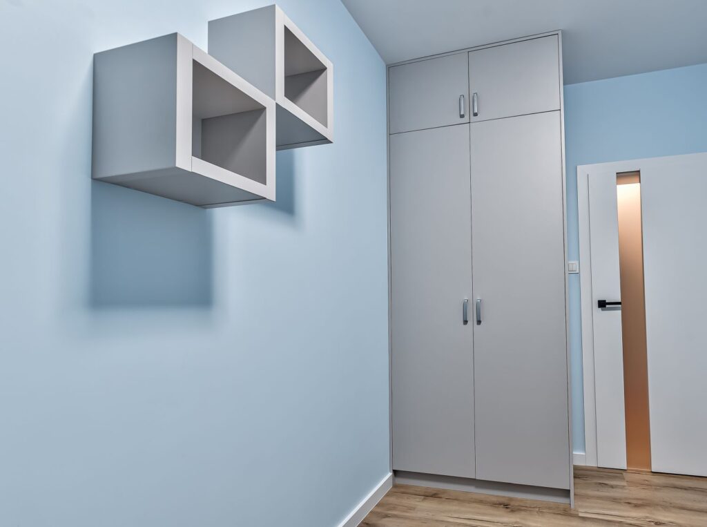 Szafka i trzy półki w minimalistycznym wnętrzu. Ściany w kolorze jasnoniebieskim, drewniana podłoga.