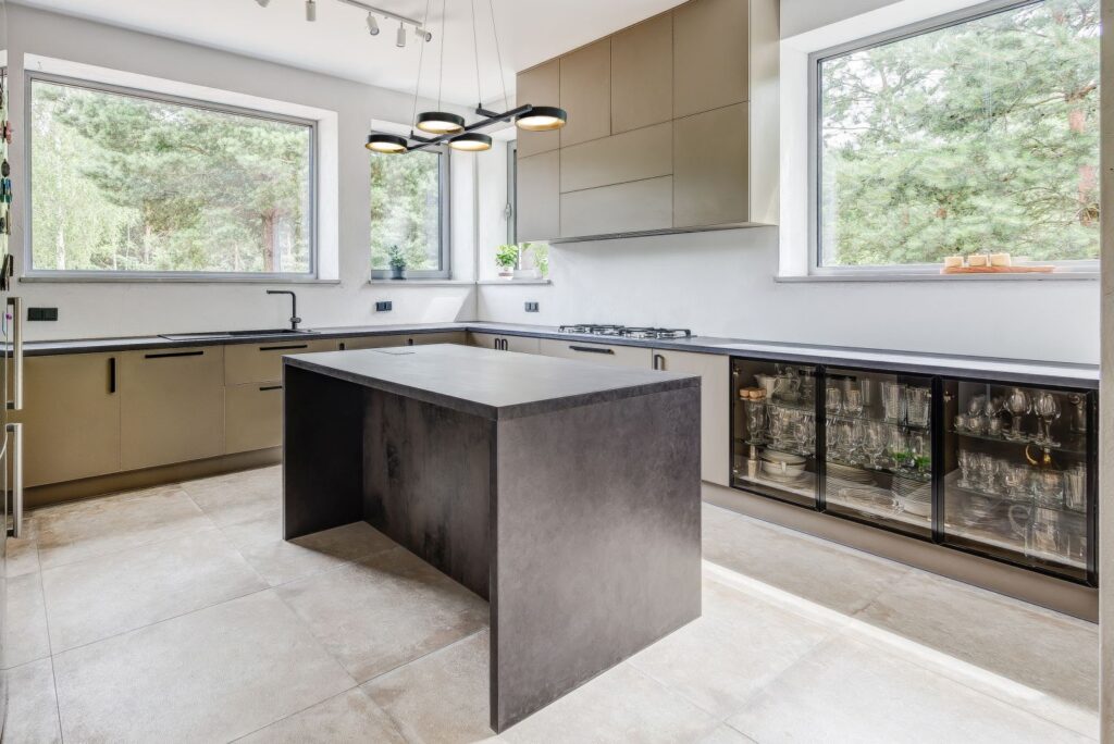 Nowoczesna kuchnia z wyspą, dużymi oknami i widokiem na las, wyposażona w czarne szafki i sprzęt AGD.
