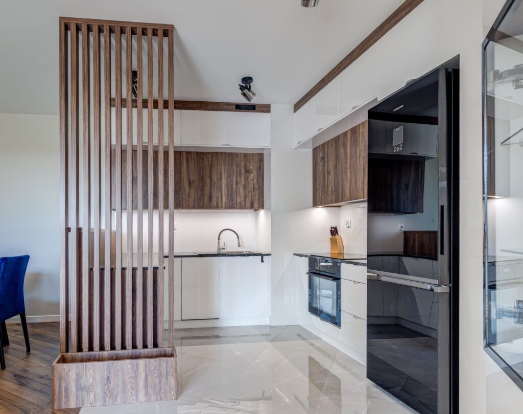 Nowoczesna kuchnia z drewnianymi i białymi szafkami, czarnym sprzętem AGD oraz drewnianą ażurową przegrodą.