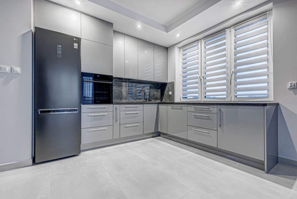 Nowoczesna kuchnia z szarymi szafkami, czarnym sprzętem AGD i dużymi oknami z żaluzjami.