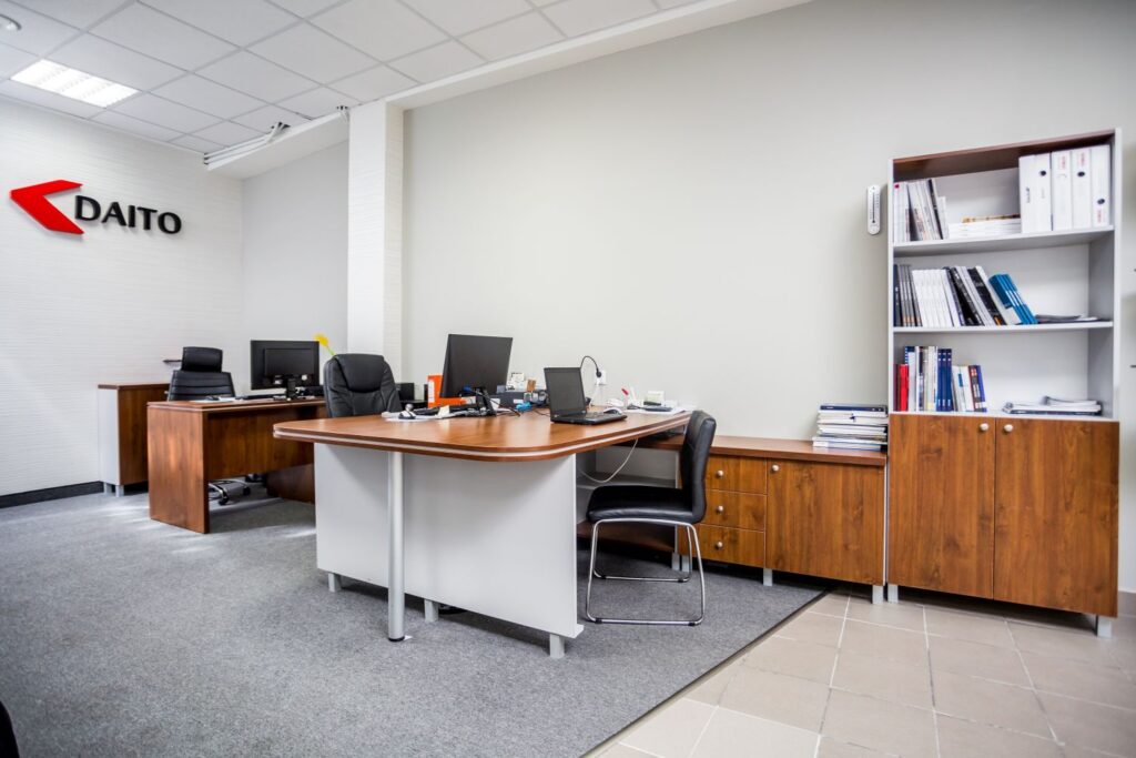 Biuro z drewnianymi biurkami, czarnymi krzesłami, komputerami i regałem pełnym książek na tle jasnej ściany.