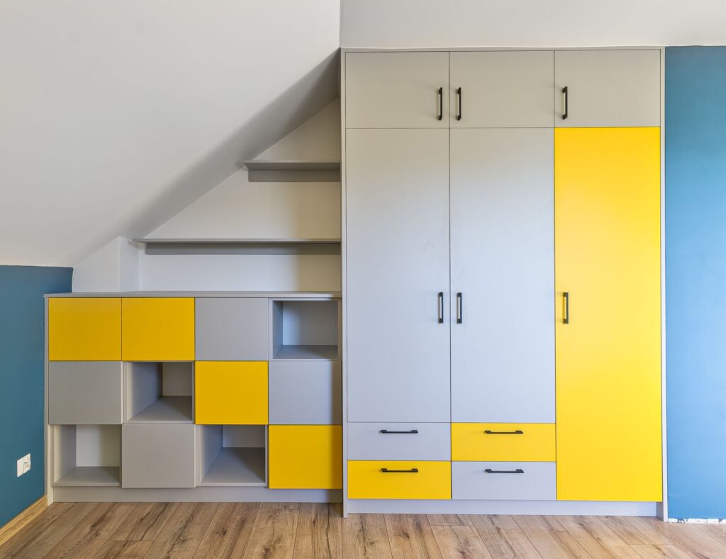 Szafka pod skosem z szarymi i żółtymi frontami, drewniana podłoga, niebieska ściana.