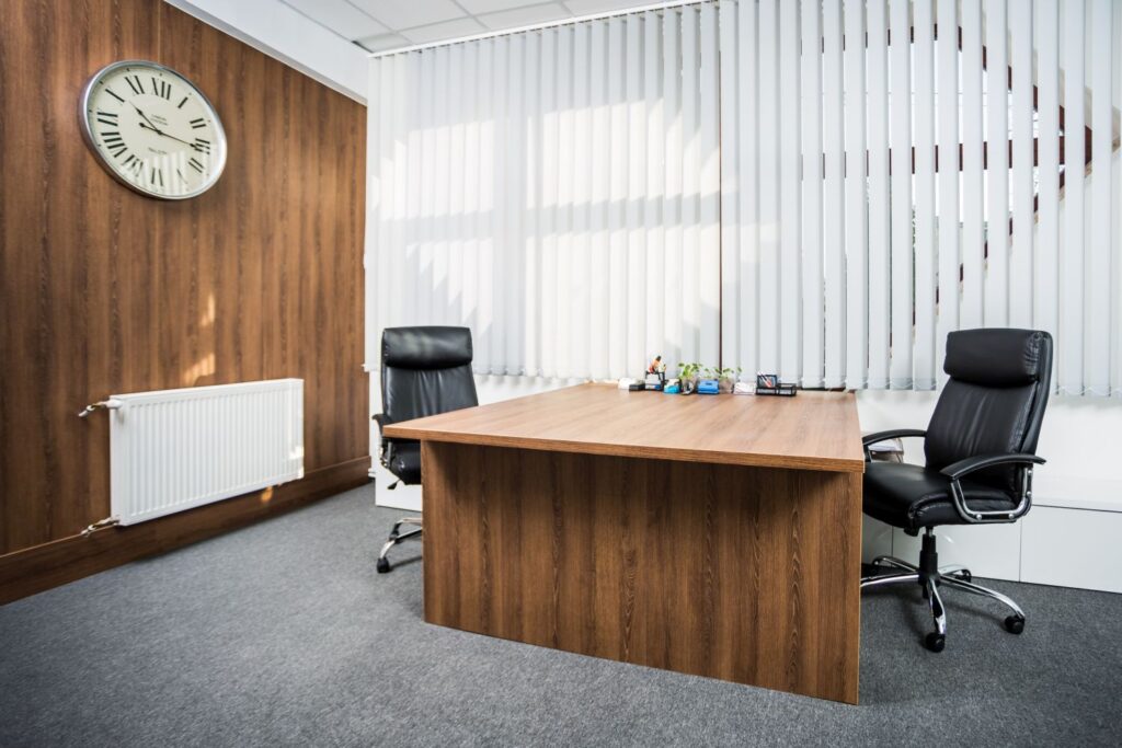 Nowoczesne biuro z drewnianym biurkiem i dwiema skórzanymi krzesłami, zegar ścienny i żaluzje w tle.
