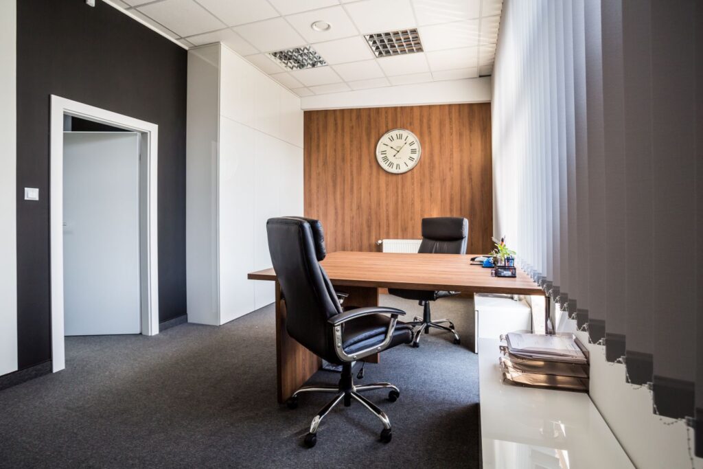 Nowoczesne biuro z drewnianym stołem, czarnymi fotelami i zegarem na ścianie. Jasne wnętrze z zasłonami pionowymi.