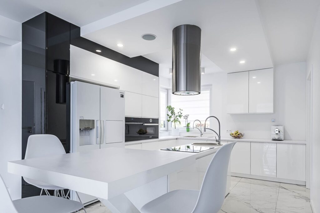 Nowoczesna biała kuchnia z wyspą, stołem, krzesłami, dużą lodówką i okapem; jasne wnętrze z roślinami na parapecie.