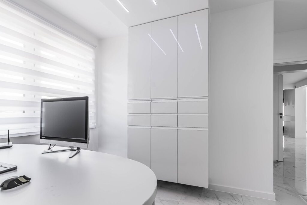 Nowoczesne, minimalistyczne biuro z białymi meblami, biurkiem, komputerem i szafą.