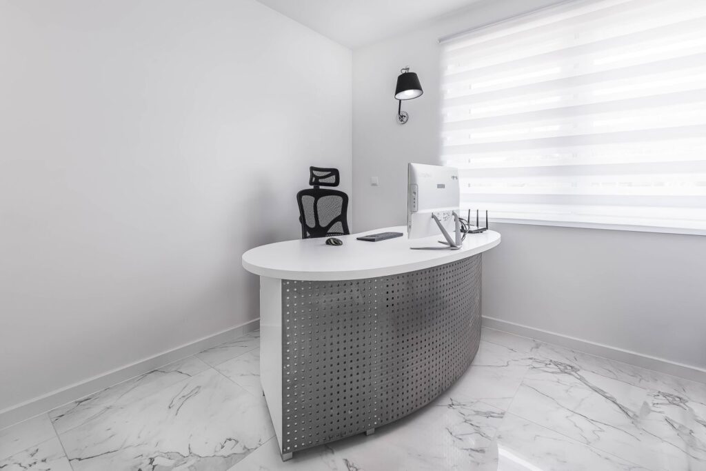 Nowoczesne biuro z białym biurkiem, krzesłem obrotowym, lampą ścienna i komputerem na marmurowej podłodze.