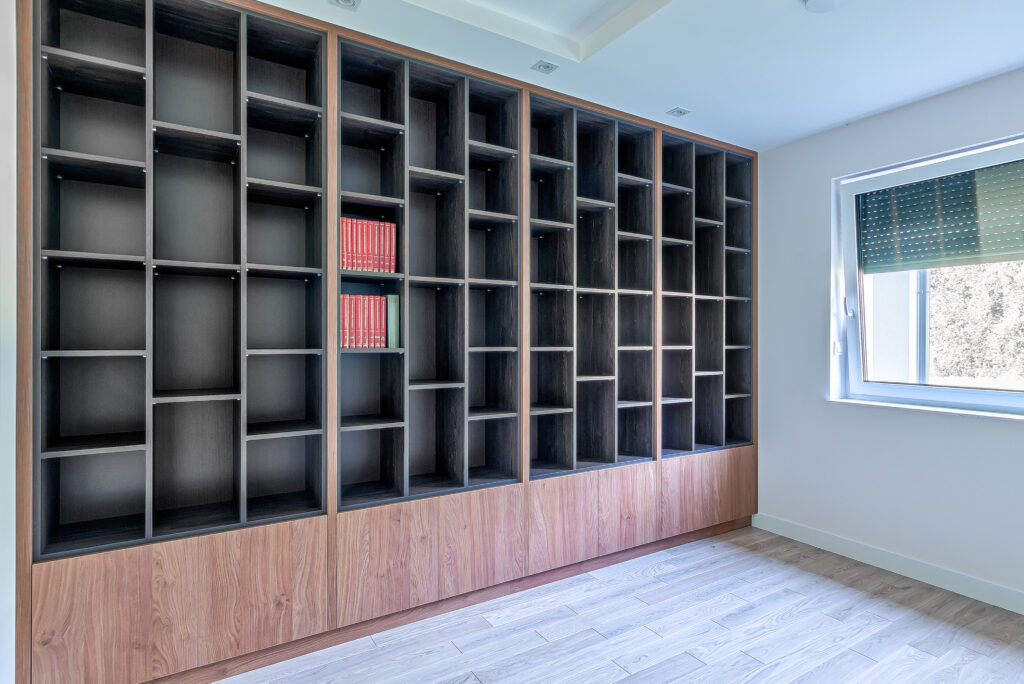 Puste regały w nowoczesnym pomieszczeniu z oknem. Na jednej półce znajdują się rzędy czerwonych książek.