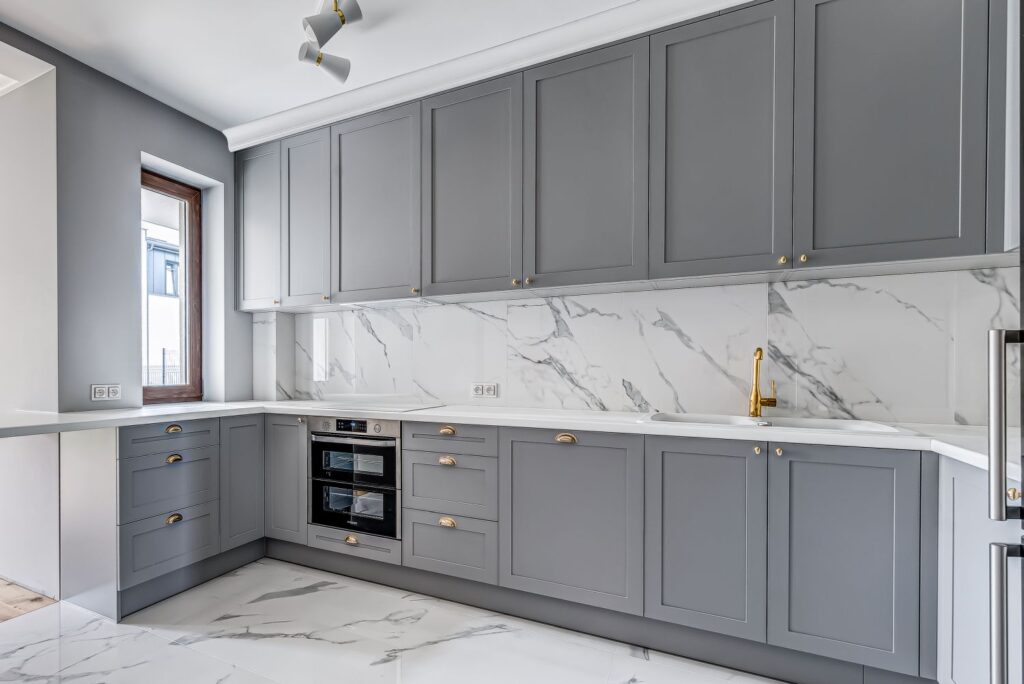 Nowoczesna kuchnia z szarymi szafkami, marmurowymi płytkami i złotym kranem. Wbudowany piekarnik i jasne wnętrze.