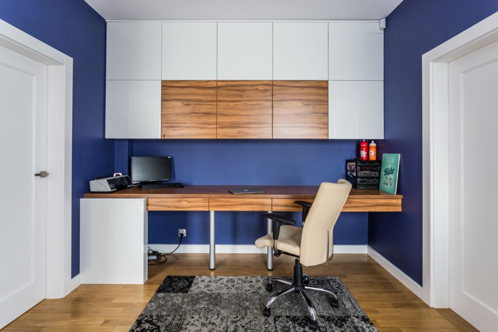 Nowoczesne biuro z jasnym biurkiem, komputerem, kremowym fotelem, szafkami i granatowymi ścianami.