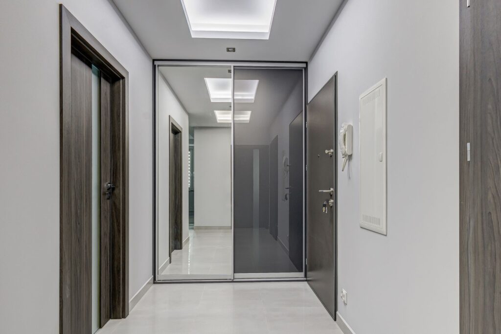 Nowoczesny korytarz z drzwiami, lustrem i szarymi ścianami, oświetlony sufitowymi panelami LED.