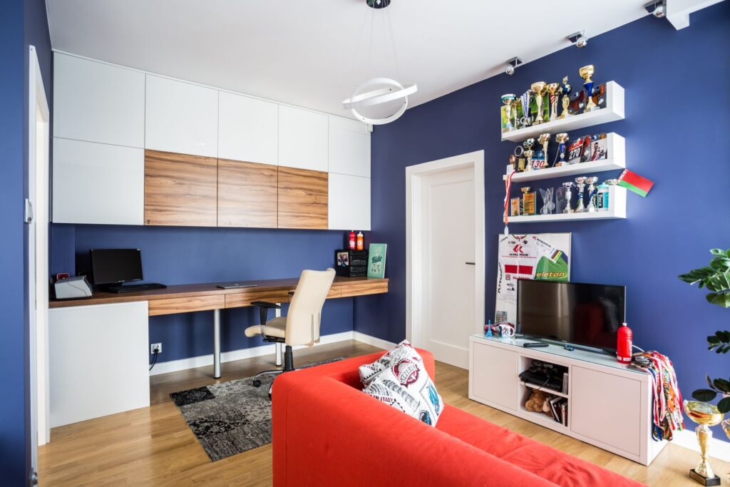 Nowoczesny pokój z biurkiem, czerwonym sofa, pucharami na półkach i niebieskimi ścianami.