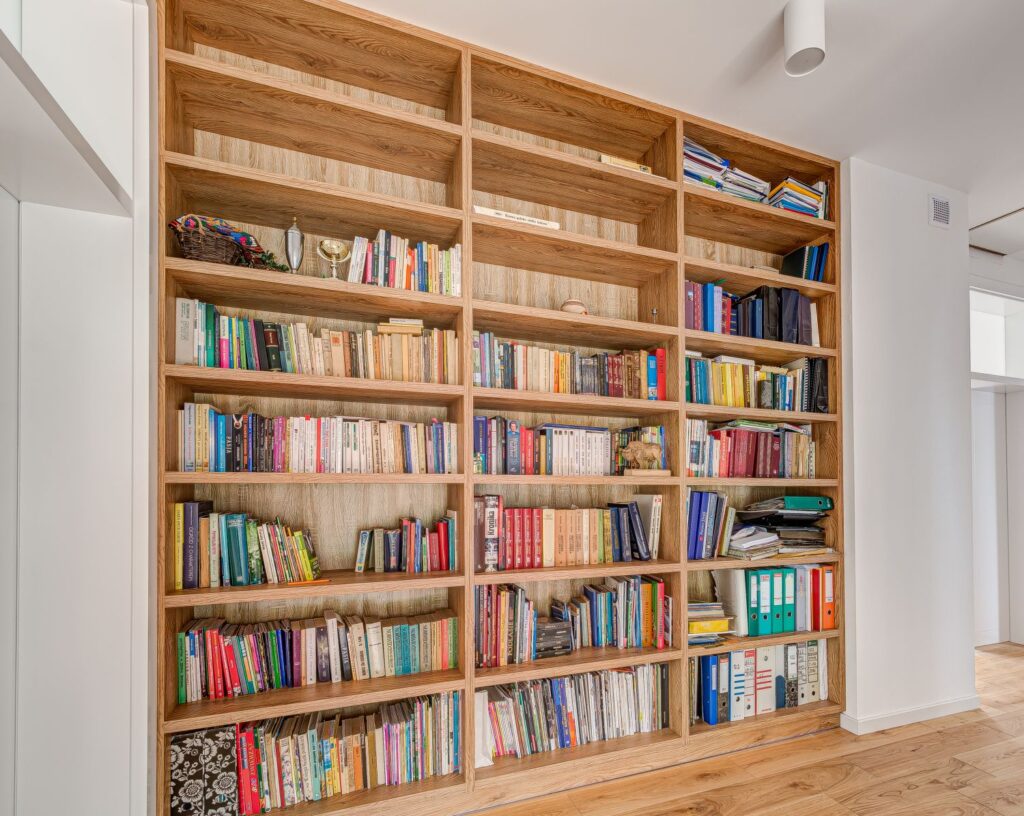Drewniana biblioteka pełna różnorodnych książek i segregatorów w nowoczesnym, jasnym wnętrzu.