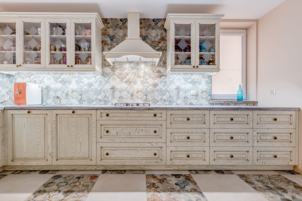 Kuchnia z jasnymi szafkami, wzorzystym backsplash'em i kwadratową podłogą. Na blacie niebieska butelka i małe urządzenie.