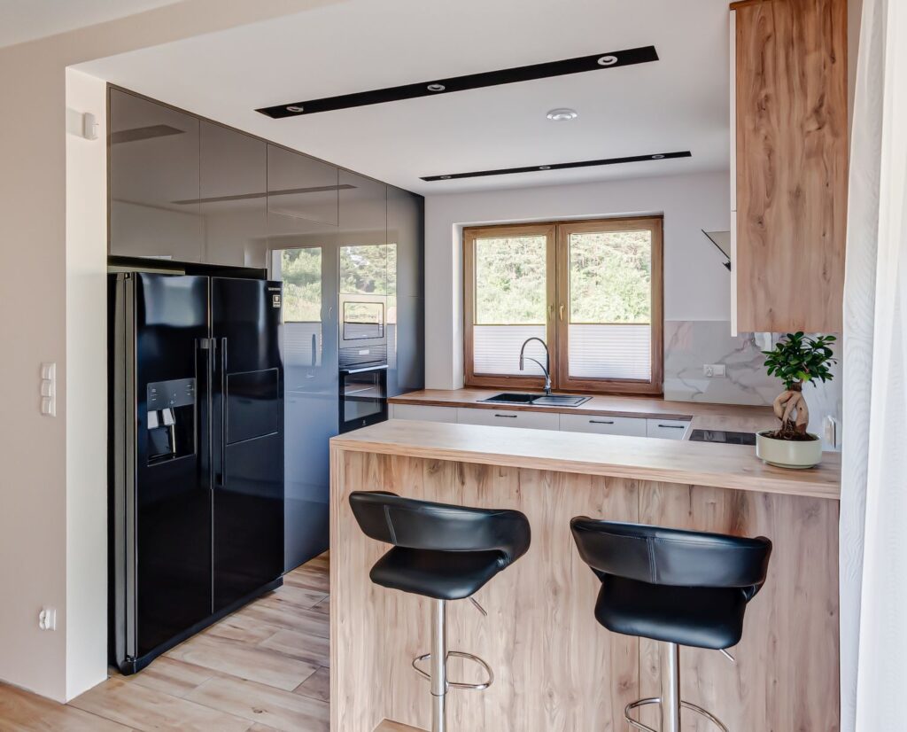 Nowoczesna kuchnia z drewnianym blatem, czarnymi krzesłami, dużą lodówką i oknem z widokiem na zieleń.
