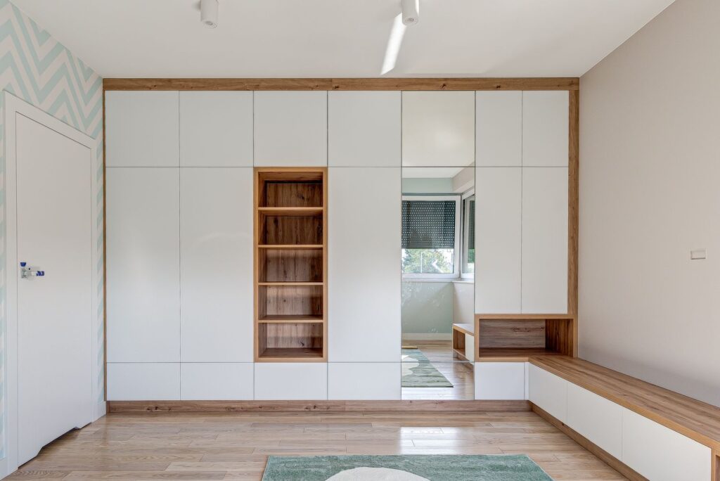 Nowoczesne białe szafy z drewnianymi akcentami w jasnym pokoju z parkietem i pasem wzorzystej tapety.