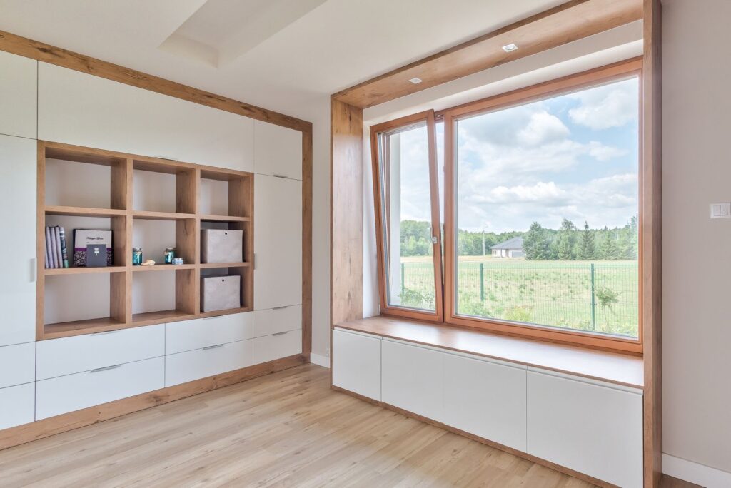 Jasny pokój z drewnianą podłogą, dużym oknem z widokiem na zieleń, nowoczesnymi półkami i szafkami.