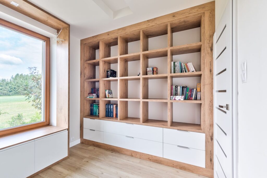 Pusta drewniana półka z książkami oraz szufladami, obok duże okno z widokiem na ogród.