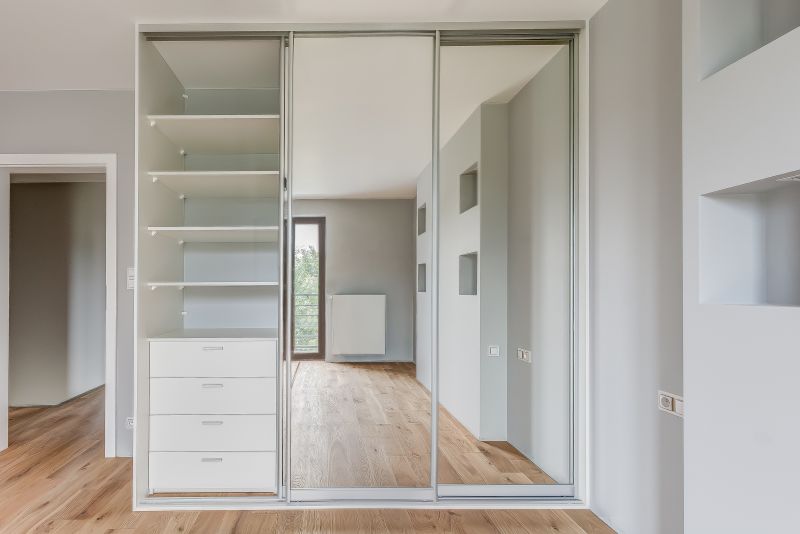 Szafa z przesuwnymi drzwiami lustrzanymi, obok otwarte półki i szuflady. Drewniana podłoga, jasne ściany.