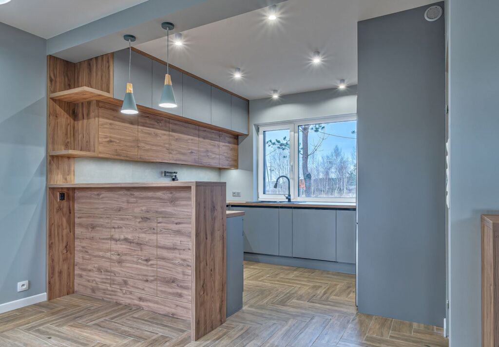 Nowoczesna kuchnia z drewnianą zabudową, szarymi szafkami, dużym oknem i wiszącymi lampami.