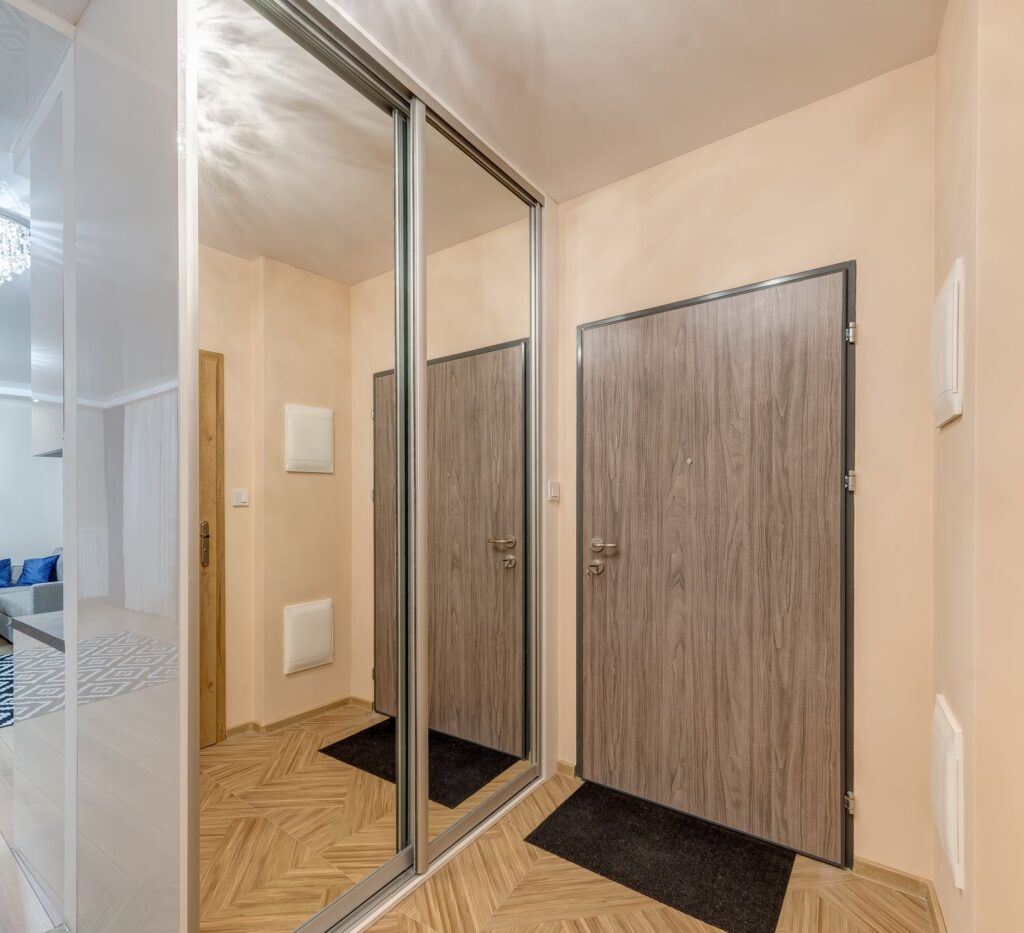 Korytarz mieszkania z drewnianymi drzwiami i lustrzaną szafą. W tle widoczny fragment salonu z dywanem i sofą.