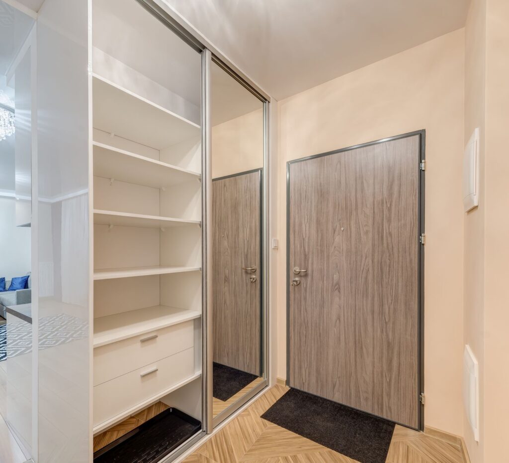 Przedpokój z lustrem, szafą wnękową z otwartymi półkami. Drzwi wejściowe w kolorze drewna. Nowoczesne, jasne wnętrze.