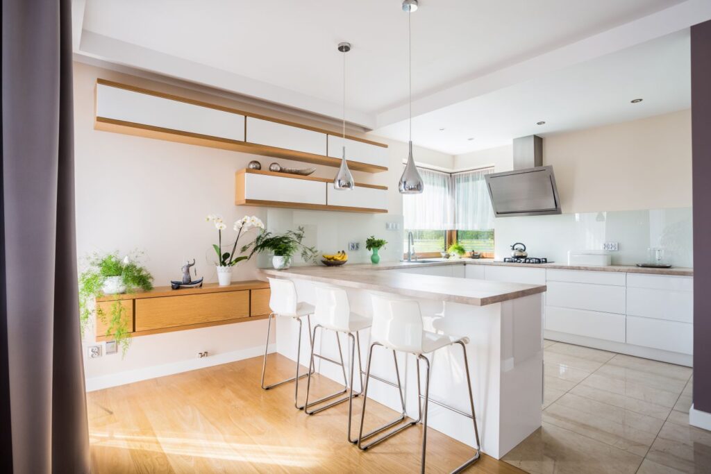 Nowoczesna, jasna kuchnia z wyspą, białymi meblami, drewnianymi akcentami i roślinami.