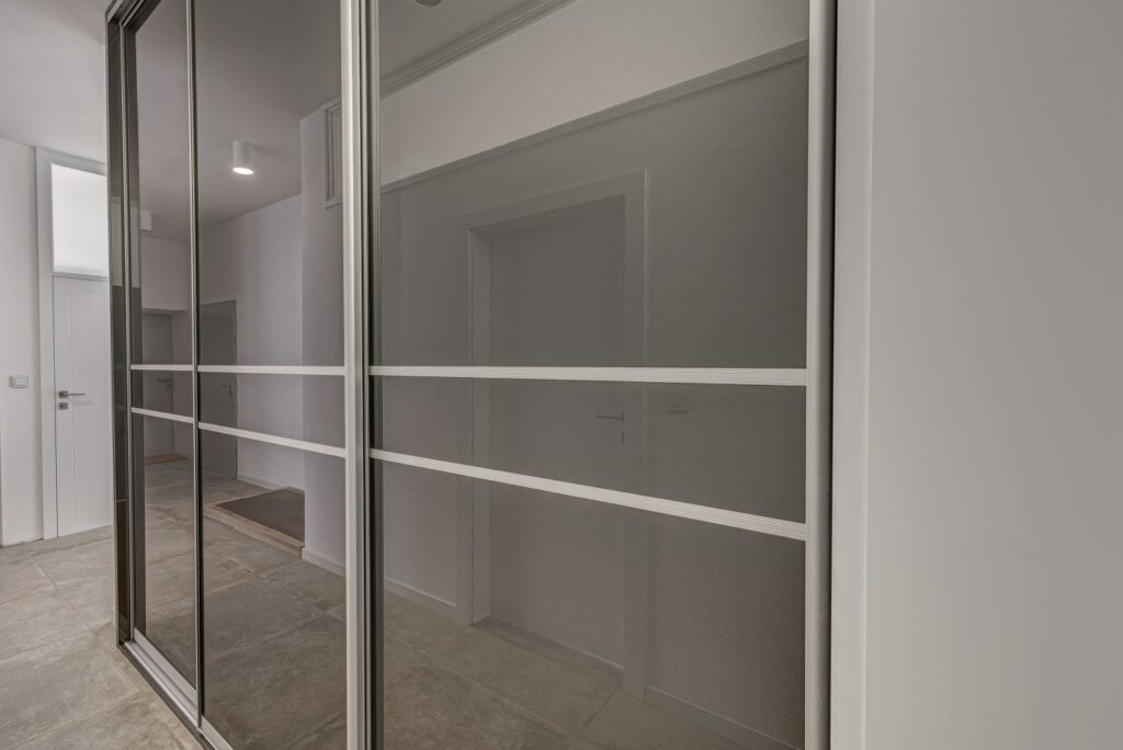Przesuwne drzwi z lustrami w nowoczesnym, jasnym wnętrzu z szarymi kafelkami na podłodze i białymi ścianami.