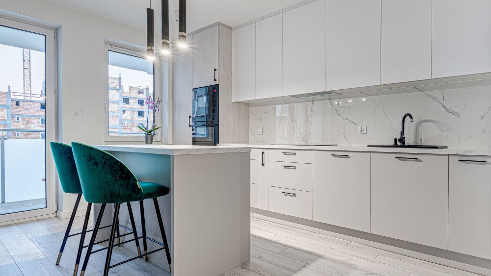 Nowoczesna kuchnia z białymi szafkami, marmurowym blatem, zielonymi krzesłami barowymi i dużymi oknami.