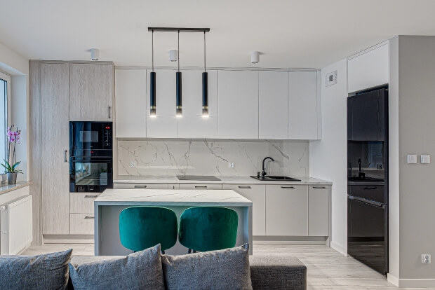 Nowoczesna kuchnia z białymi szafkami, wyspą i zielonymi krzesłami. Wiszą trzy lampy, obok stoją lodówka i piekarnik.