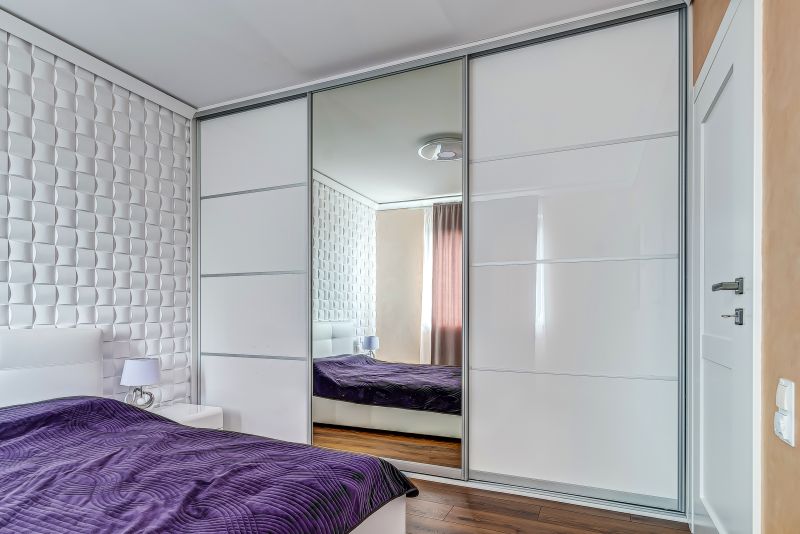 Sypialnia z białymi szafami, dużym lustrem i łóżkiem z fioletową narzutą, ściany z geometrycznym wzorem.