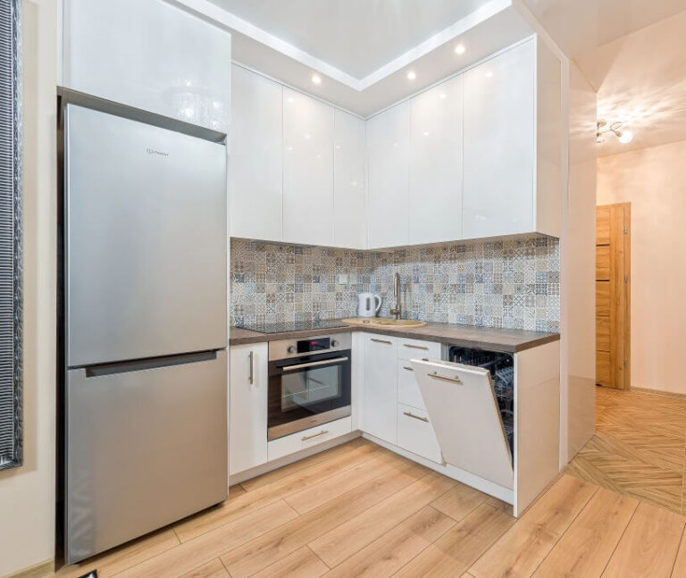 Nowoczesna kuchnia z białymi szafkami, wzorzystymi płytkami, piekarnikiem, lodówką i zmywarką w zabudowie.