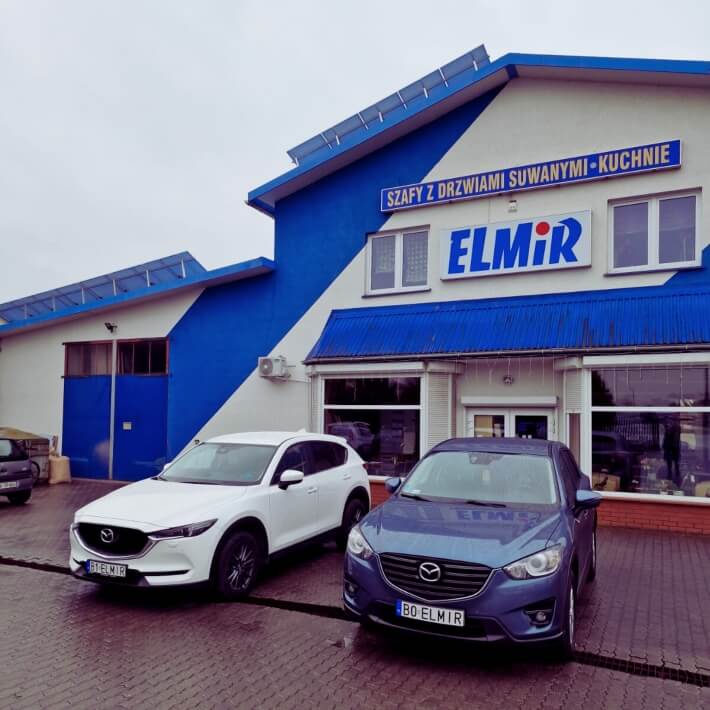 Budynek sklepu Elmir z niebieskim dachem i dwoma samochodami Mazda zaparkowanymi przed wejściem.
