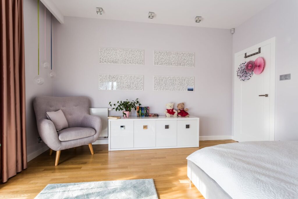 Jasny pokój z fotelem, komodą i dekoracjami. Na komodzie stoi roślina i siedzą pluszowe misie.