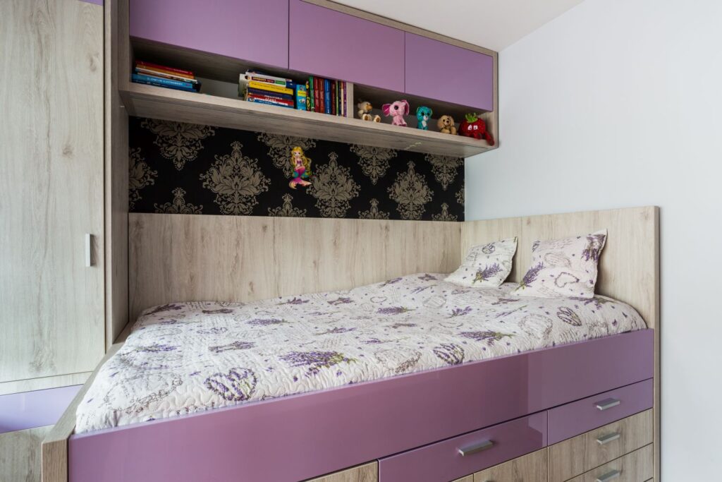 Łóżko z fioletowymi szufladami, nad nim półka z książkami i maskotkami, ozdobna tapeta w tle.