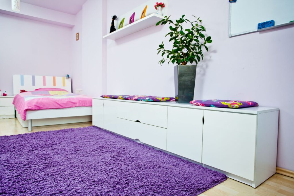 Pokój dziecięcy z różowym łóżkiem, białymi meblami, fioletowym dywanem i rośliną w doniczce.