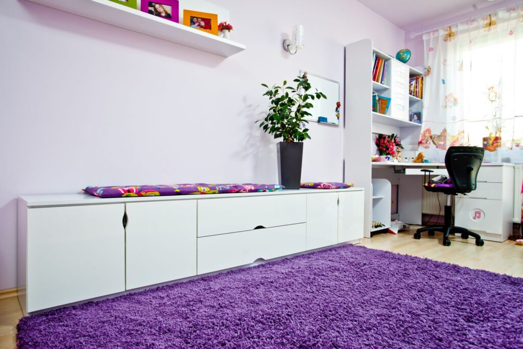 Jasny pokój dziecięcy z białymi meblami, fioletowym dywanem, rośliną w doniczce i biurkiem przy oknie.
