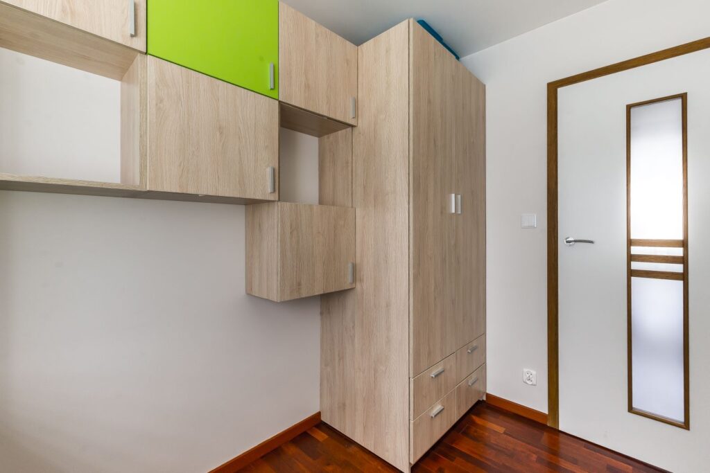 Szafki i szafa z jasnego drewna, jedna z zielonym frontem, w nowoczesnym wnętrzu z drewnianą podłogą.