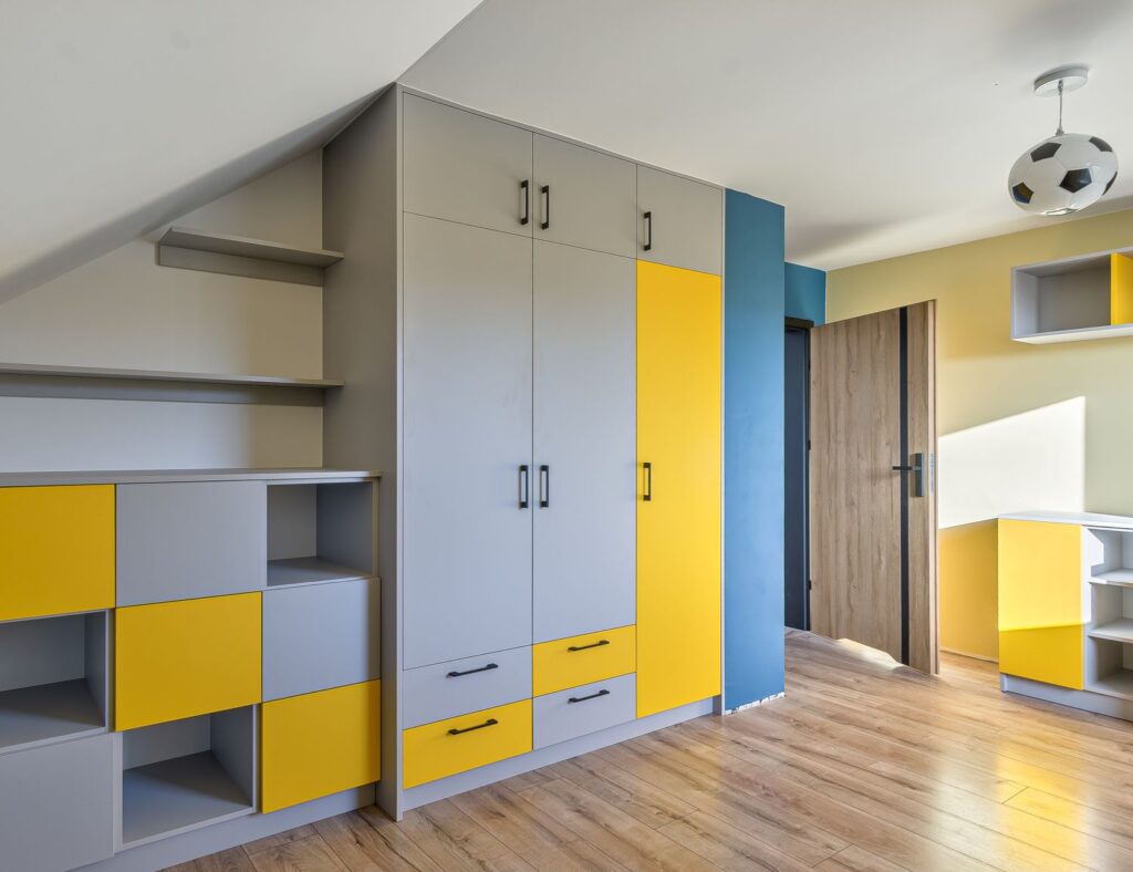 Pokój w jasnych kolorach, z żółtymi i szarymi meblami, półkami oraz lampą w kształcie piłki.