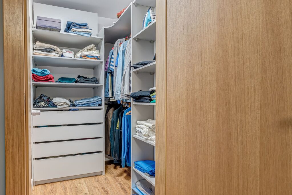 Przestronna garderoba z otwartymi półkami pełnymi kolorowych ubrań, koszulami i spodniami wiszącymi na wieszakach.