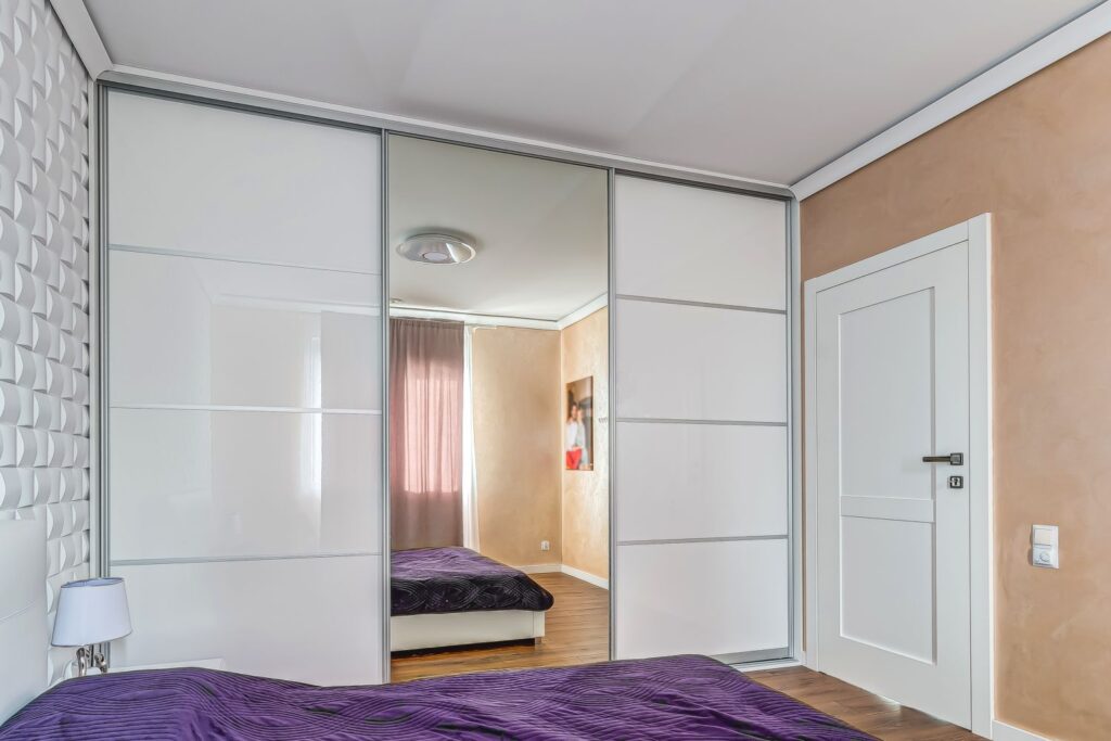 Nowoczesna sypialnia z lustrzaną szafą, fioletową pościelą, jasnymi ścianami i dużym białym drzwiami.