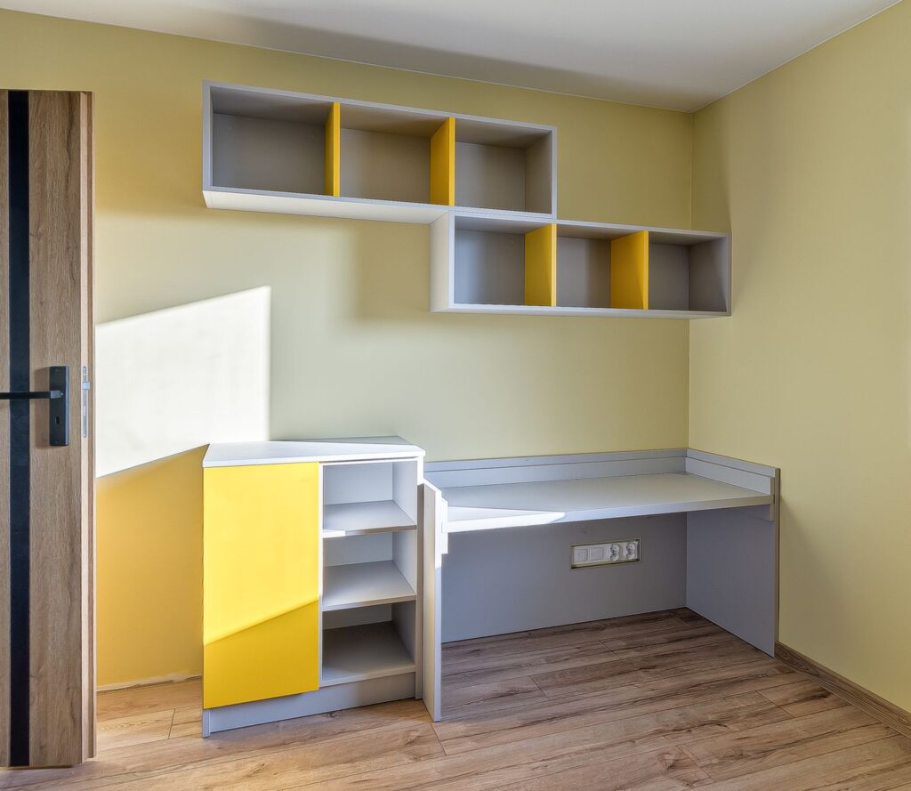 Pokój z żółtymi i szarymi półkami, biurkiem oraz drewnianymi podłogami. Jasne, pastelowe ściany dodają przestrzeni.