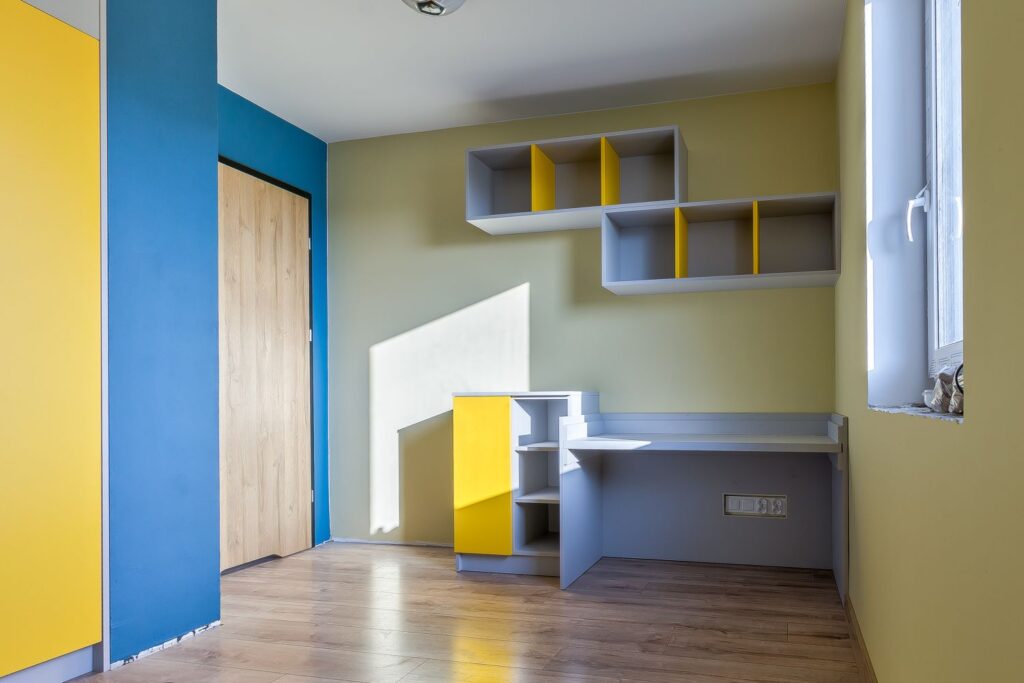 Pokój z jasnymi ścianami, kolorowym biurkiem i półkami, przytulnie oświetlony słońcem wpadającym przez okno.
