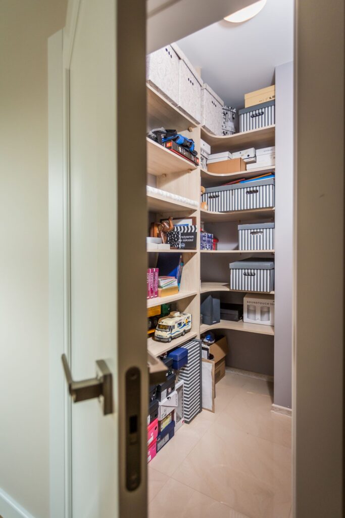 Otwarta szafa z półkami pełnymi pudełek i różnych przedmiotów, widok przez niedomknięte drzwi.