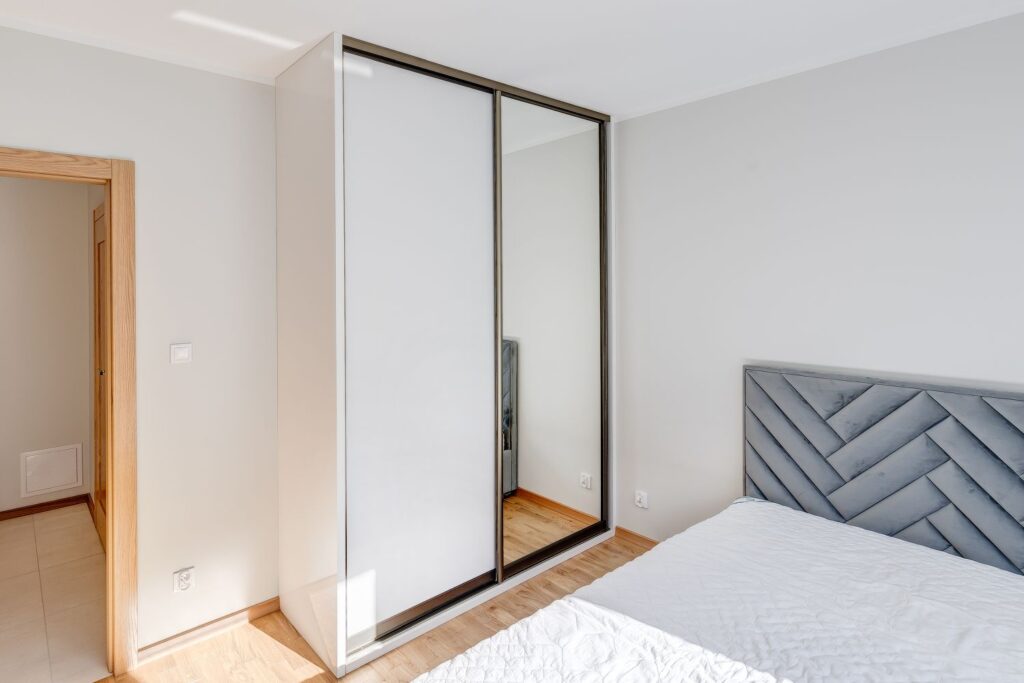 Nowoczesna sypialnia z jasnymi ścianami, dużą szafą z lustrzanymi drzwiami i łóżkiem z szarym zagłówkiem.